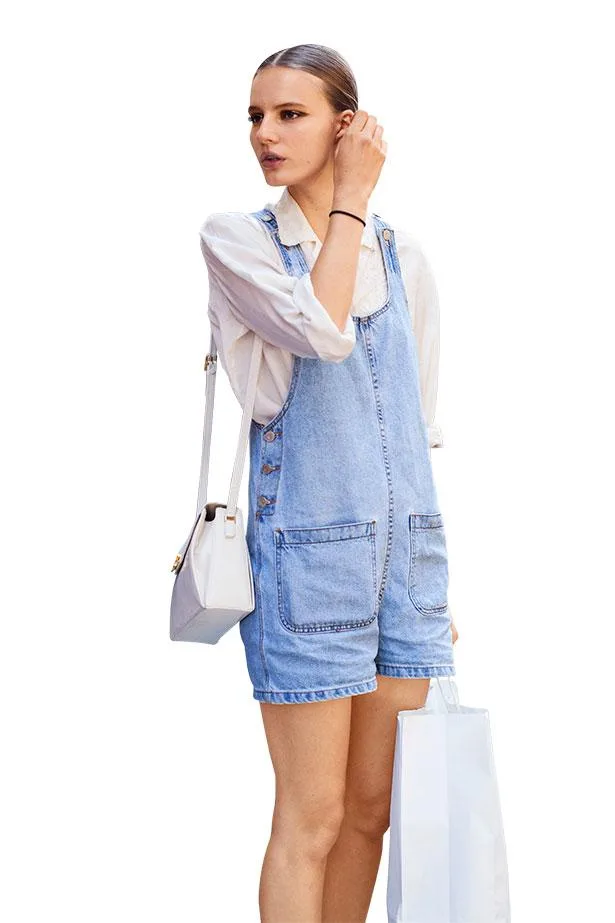 A woman in a white shirt and denim overalls, holding a shopping bag and a white handbag.