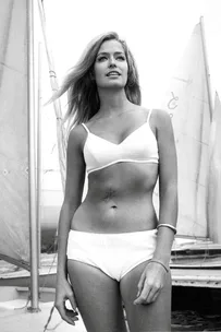 A woman in a white bikini standing on a boat with sails in the background, smiling and looking forward.