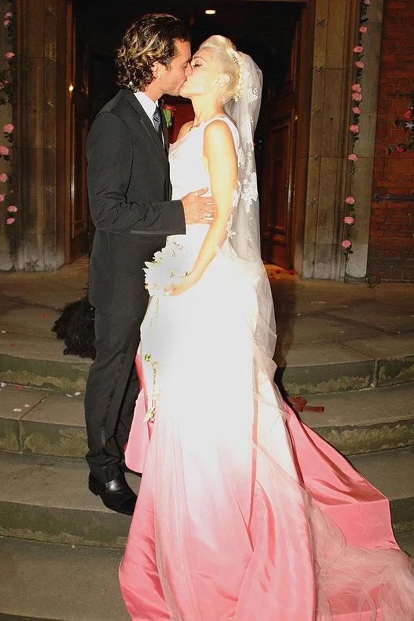 A bride in a pink gown kisses a groom outside a building, both in formal attire.