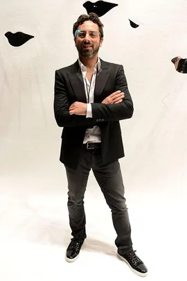 Man in a black blazer and jeans wearing smart glasses, crossing his arms and smiling.