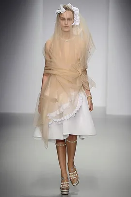 Model on runway in a layered beige veil, white dress, flower sandals, and headpiece, showcasing a bridal look.