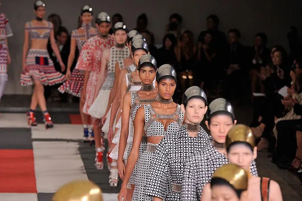 Fashion models in futuristic outfits and helmets walk down a runway at a fashion show.