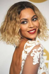 Woman with wavy blonde hair in a white lace dress smiling over her shoulder.