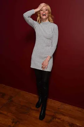 Woman in a black and white checkered dress, laughing with one hand on her head, against a maroon wall.