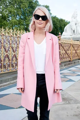 Person in a pink coat and sunglasses stands in front of ornate fence and statue in daylight.