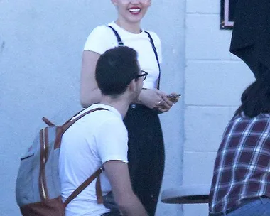 A person smiling in a white shirt with suspenders, short blonde haircut, standing outside near seated individuals.