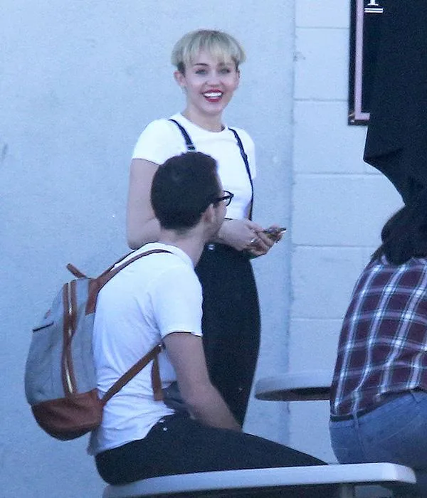 A person smiling in a white shirt with suspenders, short blonde haircut, standing outside near seated individuals.