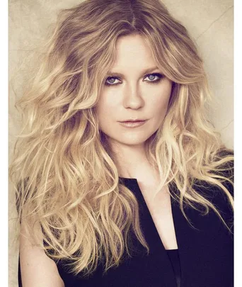 Portrait of Kirsten Dunst with voluminous, wavy blonde hair, wearing a black top against a beige background.