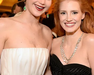 Two women in elegant dresses smiling at a formal event under a chandelier.