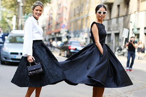 Two women smiling in elegant street fashion, wearing flared skirts and sunglasses, with a city backdrop.