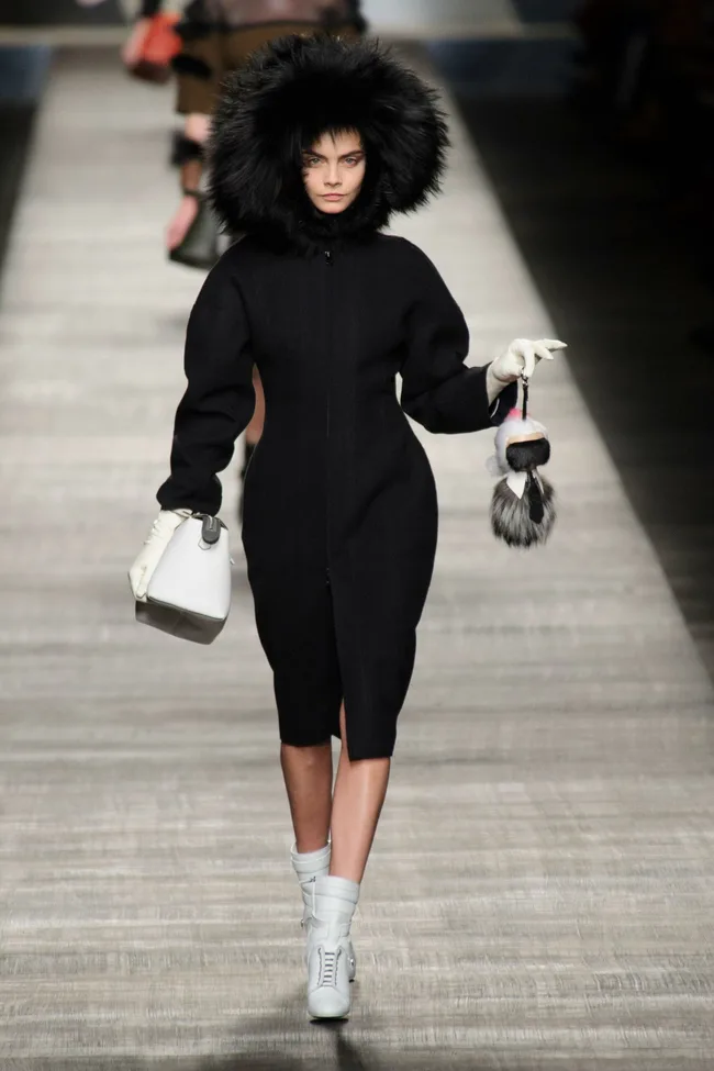 Model wearing black fur coat and hood, white gloves, and holding a handbag, on the Fendi AW14 runway.