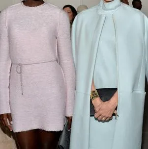 Two women pose in stylish pastel outfits; one wears a pale pink dress, the other a light blue coat, at a fashion event.