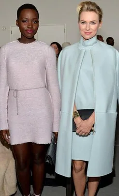 Two women pose in stylish pastel outfits; one wears a pale pink dress, the other a light blue coat, at a fashion event.