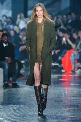 Model in olive green coat and dress with high boots walks runway. Audience in background.