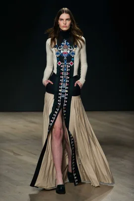 Model walking runway in a long dress with intricate patterns and a high slit, hands in pockets, on a wooden floor.