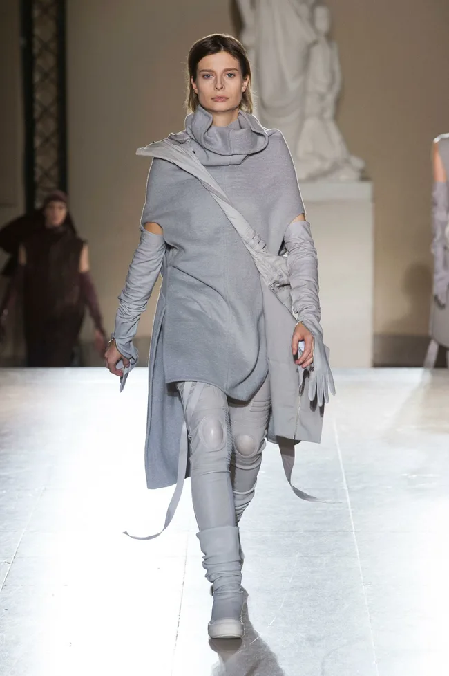 Model walking in a gray, layered outfit with high boots on a runway.