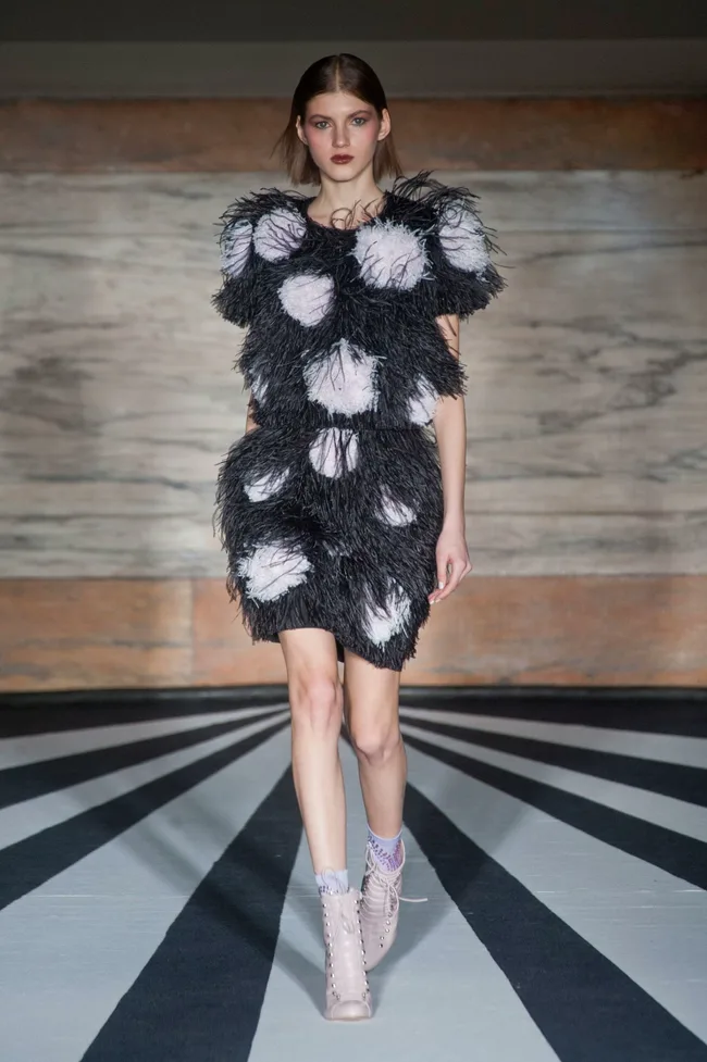 Model in black and white textured dress walks runway, background features striped pattern.