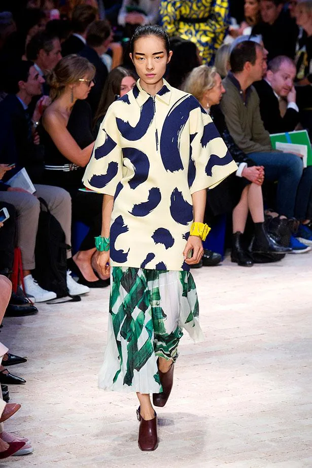 Model walking runway in a bold, abstract print dress, with a green and white pleated skirt. Audience watches in background.