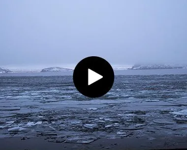 A frozen lake with patches of ice and distant snow-covered islands under a gray, cloudy sky.