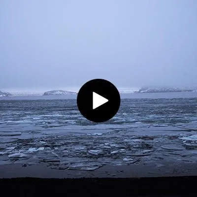 A frozen lake with patches of ice and distant snow-covered islands under a gray, cloudy sky.