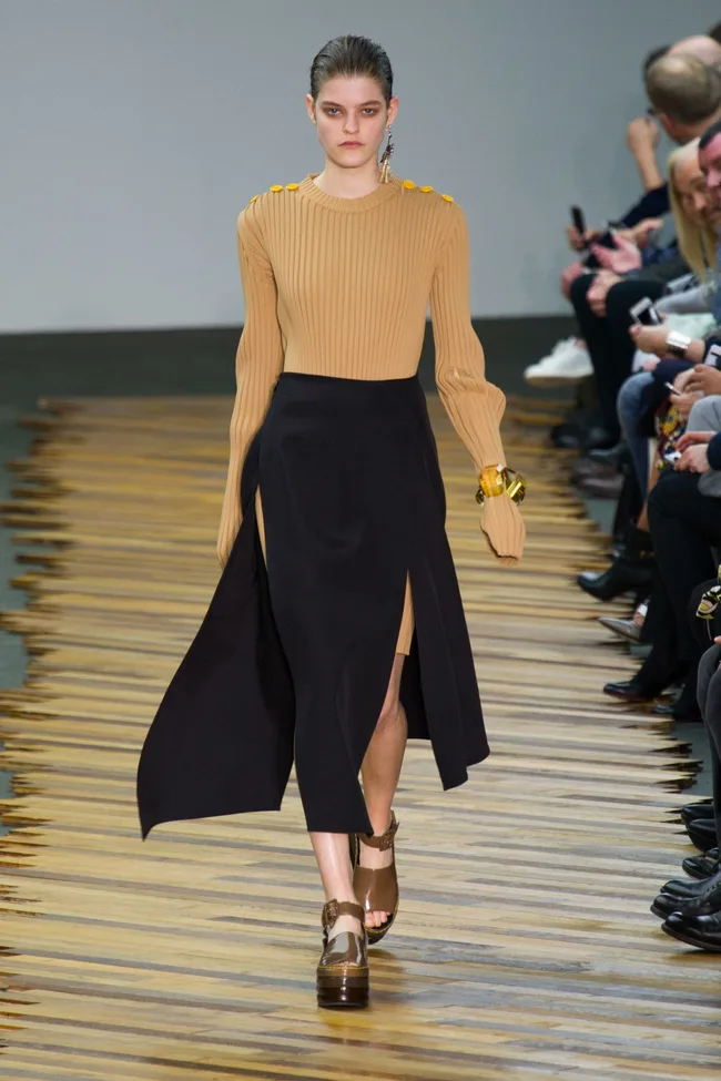 Model in beige sweater, black skirt, and platform sandals walking on a wooden runway during a fashion show.