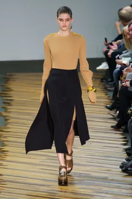 Model in beige sweater, black skirt, and platform sandals walking on a wooden runway during a fashion show.