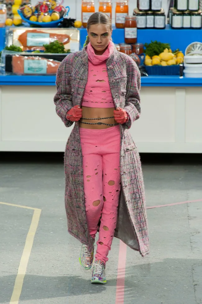 Model in pink, distressed outfit with long tweed coat and sneakers, walking on Chanel AW14/15 runway; grocery store backdrop.