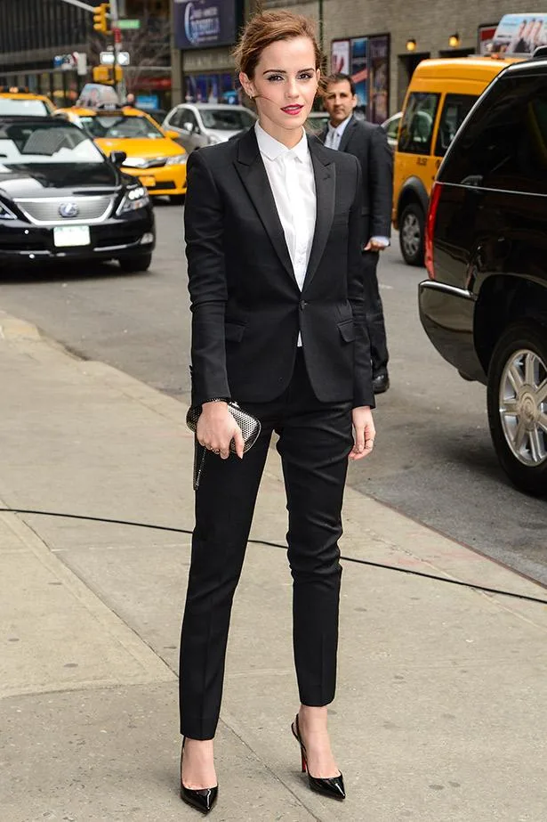 A woman in a black suit with a white shirt, holding a clutch purse, stands on a city street with taxis in the background.