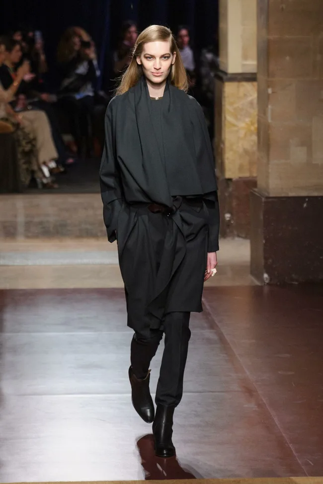 Model walks runway in a dark, elegant outfit and boots at a fashion show with audience in the background.