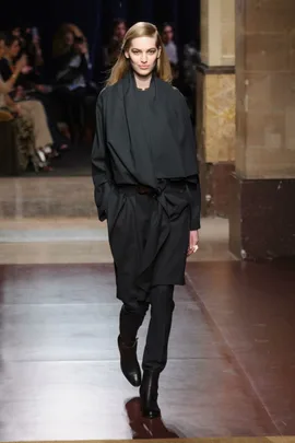 Model walks runway in a dark, elegant outfit and boots at a fashion show with audience in the background.