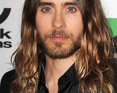 Long-haired man with a beard in a black suit, posing at an event with a stylish, natural look.