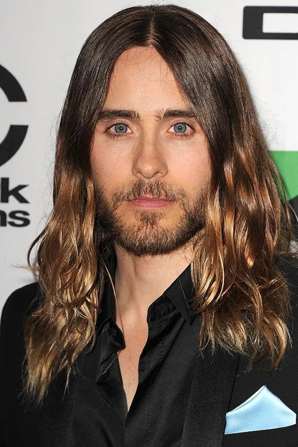 Long-haired man with a beard in a black suit, posing at an event with a stylish, natural look.