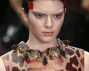 Model wearing an animal print top, intricate necklace, and red face tape on the side of eyebrows on a fashion runway.