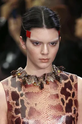 Model wearing an animal print top, intricate necklace, and red face tape on the side of eyebrows on a fashion runway.