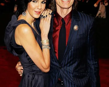 A man and woman pose together on a red carpet, both smiling and dressed in formal attire, surrounded by photographers.