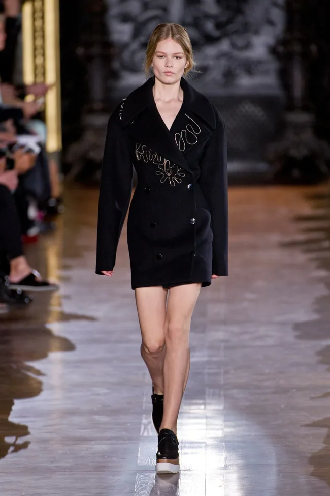 Model in a black coat with embroidery, walking on a wooden runway during Stella McCartney's AW14-15 fashion show.