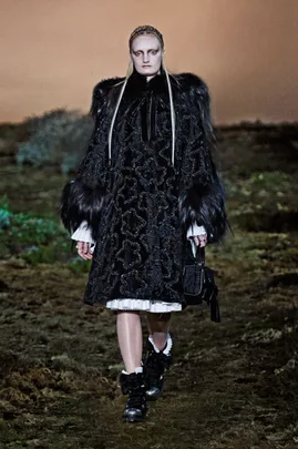 Model in a black textured coat with fur trim walks a runway, wearing dark boots and carrying a small black handbag.