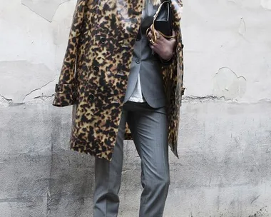 Blonde woman in leopard coat, gray suit, holding clutch, wearing Adidas sneakers and sunglasses, standing against a wall.