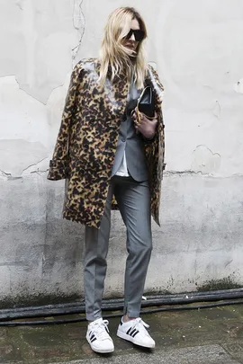 Blonde woman in leopard coat, gray suit, holding clutch, wearing Adidas sneakers and sunglasses, standing against a wall.