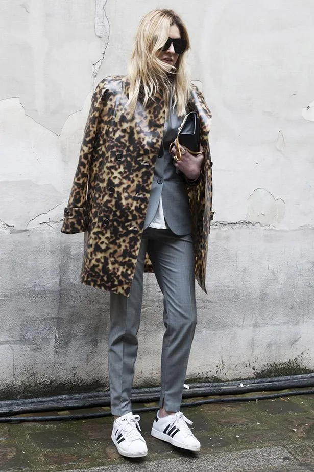 Blonde woman in leopard coat, gray suit, holding clutch, wearing Adidas sneakers and sunglasses, standing against a wall.