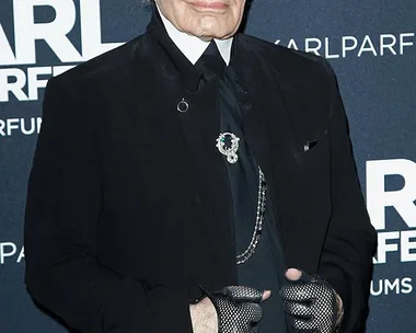 Karl Lagerfeld at perfume launch event in Paris, wearing a black outfit with sunglasses and gloves. Text on backdrop: "KARL LAGERFELD PARFUMS".