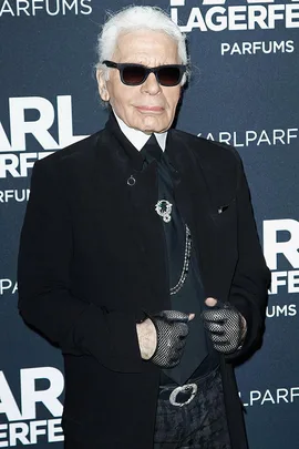 Karl Lagerfeld at perfume launch event in Paris, wearing a black outfit with sunglasses and gloves. Text on backdrop: "KARL LAGERFELD PARFUMS".