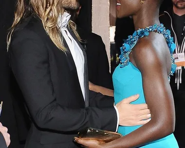 Two individuals in formal attire warmly greet each other at an event, one in a black tuxedo and the other in a turquoise gown.