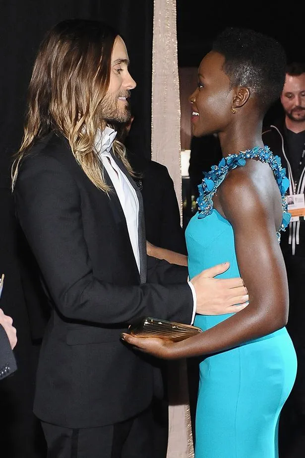 Two individuals in formal attire warmly greet each other at an event, one in a black tuxedo and the other in a turquoise gown.