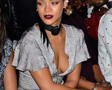 Stylish woman sitting in a gray dress with dark lipstick and a choker, surrounded by people at a fashion event.