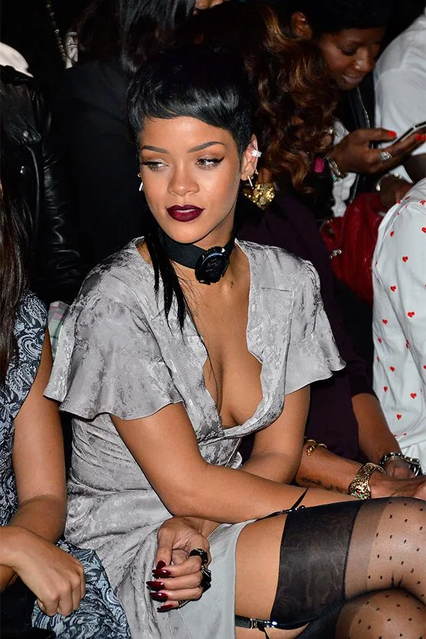 Stylish woman sitting in a gray dress with dark lipstick and a choker, surrounded by people at a fashion event.