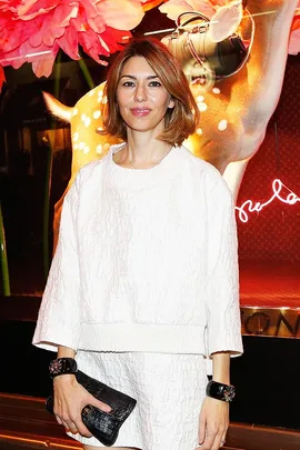 A woman in a white outfit holding a black clutch, standing in front of a display with large flowers and a deer figure.