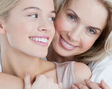 A mother and daughter embracing and smiling at each other with a gray background.