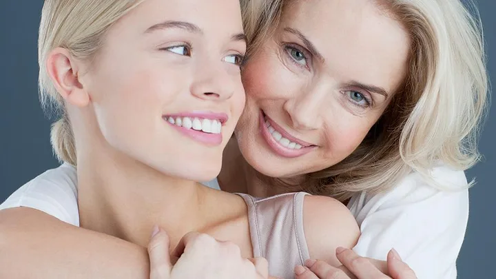 A mother and daughter embracing and smiling at each other with a gray background.