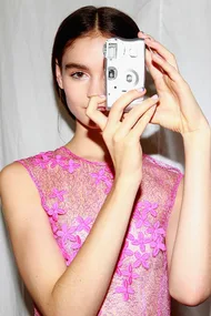 Model in a pink lace dress holding a vintage camera, posing at fashion week.